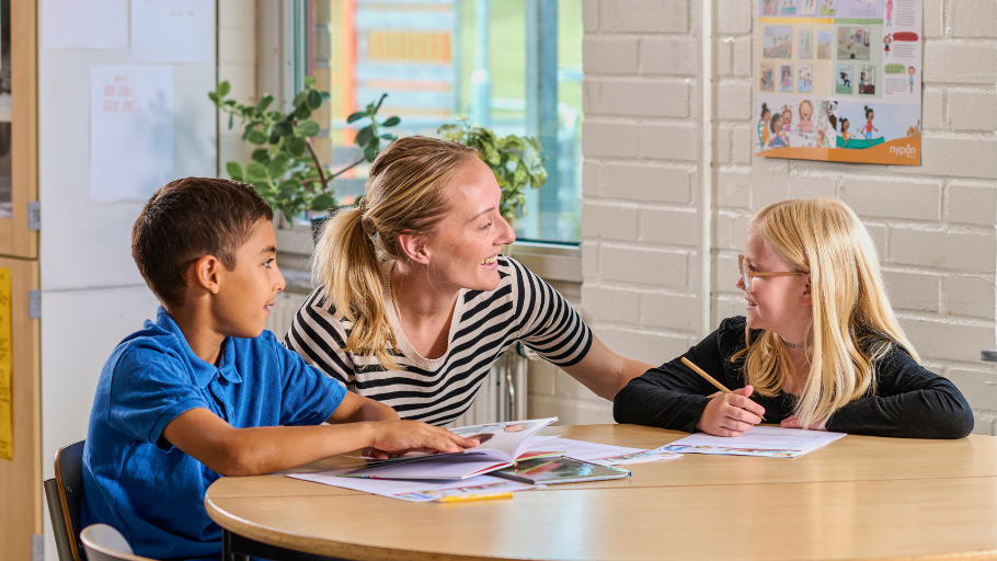 Kvinna i skolmiljö sitter mellan två elever, en pojke och en flicka, som skriver och läser böcker. De ser glada ut.
